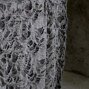May include: Gray tattered fabric with a distressed, ripped, and torn texture. The fabric is draped over a surface and has a lace-like appearance.