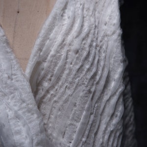 May include: White, textured fabric with a loose weave, creating a flowing, draped effect. The fabric is soft and delicate, with a slightly sheer appearance.