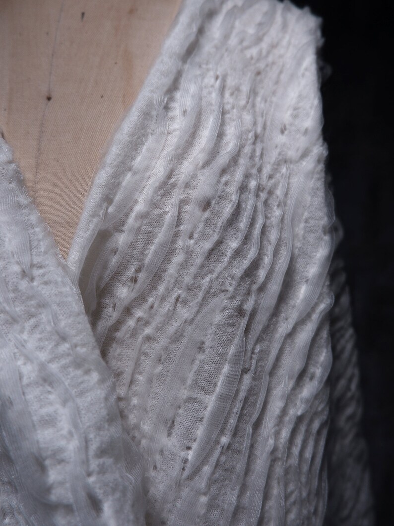 May include: White, textured fabric with a loose weave, creating a flowing, draped effect. The fabric is soft and delicate, with a slightly sheer appearance.