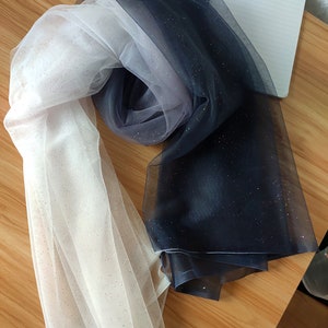 Peut inclure: Un tissu de tulle ombré blanc et bleu marine avec des paillettes. Le tissu est transparent et a une texture douce et fluide.