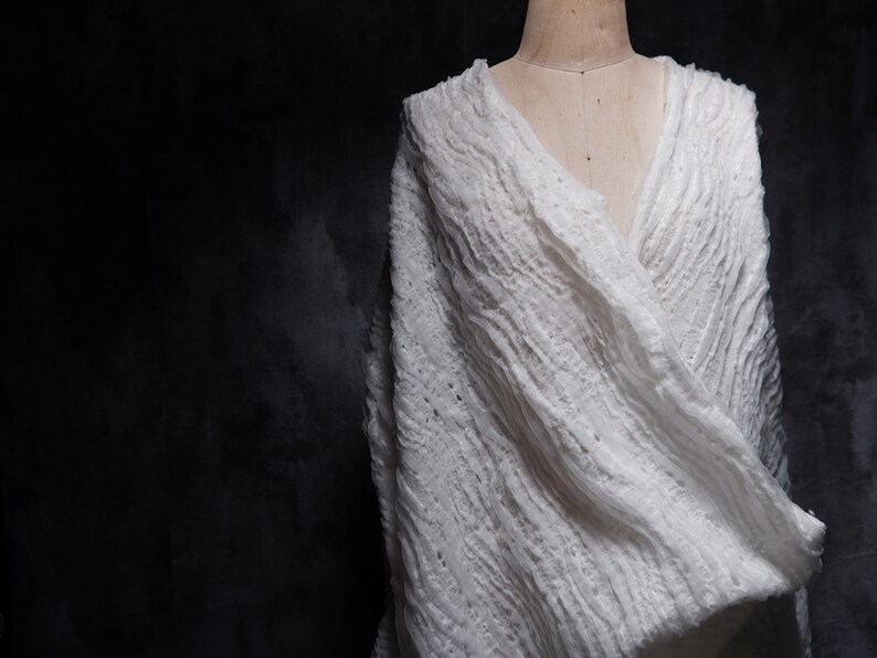 May include: A white, textured, crocheted shawl draped over a mannequin. The shawl has a loose, flowing design and is made of a soft, lightweight fabric.
