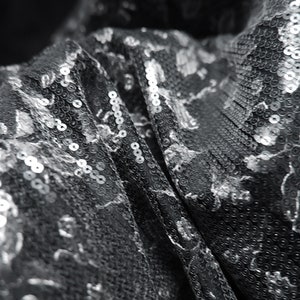 May include: Close-up of a silver sequined fabric with a textured pattern. The fabric is folded and the sequins are catching the light.