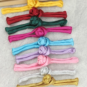 Könnte beinhalten: Sortiment aus zehn bunten Stoff-Chinesischen Knotenverschlüssen. Die Knoten sind in Blumenform gebunden und in Gelb, Rot, Grün, Rosa, Blau, Lila, Silber und Gold erhältlich.