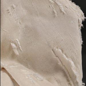 May include: Close-up of a piece of off-white fabric with a textured weave. The fabric has several frayed edges and tears.