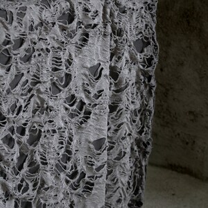 May include: Gray tattered fabric with a distressed, ripped, and torn texture. The fabric is draped over a wooden surface.