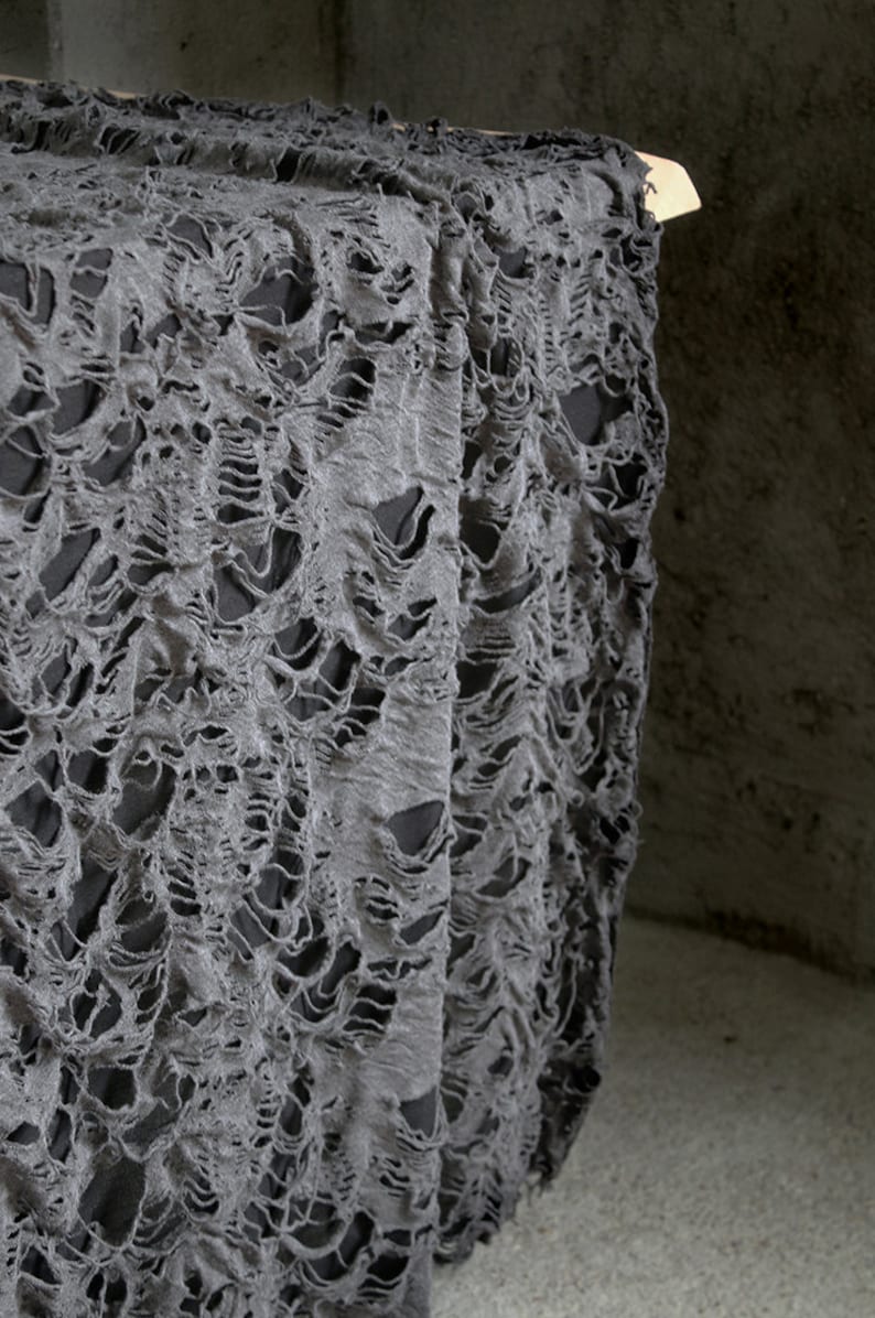 May include: Gray tattered fabric with a distressed, ripped, and torn texture. The fabric is draped over a wooden surface.