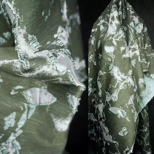 Puede incluir: Primer plano de una tela drapeada con un patrón de formas blancas y verde claro sobre un fondo verde oliva oscuro. La tela tiene una apariencia texturizada y está doblada, revelando el reverso de color verde claro.