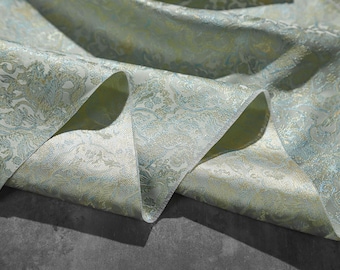 May include: A close-up of a light green and gold fabric with a paisley pattern. The fabric is folded and draped, showing the texture and detail of the design.