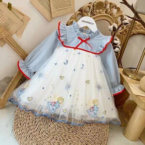 Puede incluir: Un vestido de tul blanco con mangas de mezclilla azul y una cinta roja. El vestido est&aacute; decorado con estrellas, lunas y &aacute;ngeles bordados. El vestido tiene un cuello con volantes y una etiqueta que dice "Mimibello".