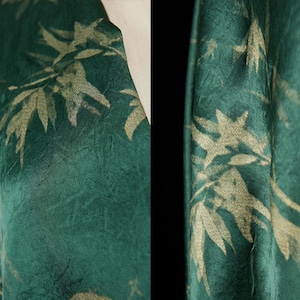 May include: Close-up of a teal fabric with a gold bamboo leaf pattern. The fabric has a textured appearance and is draped, revealing the pattern's intricate details. The design is repeated across the textile.