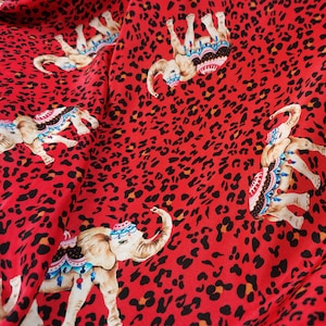 May include: Red fabric with a repeating pattern of black leopard spots and colorful elephants with ornate decorations. The elephants are facing different directions.
