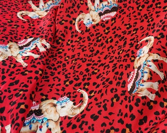 May include: Red fabric with a repeating pattern of black leopard spots and colorful elephants with ornate decorations. The elephants are facing different directions.