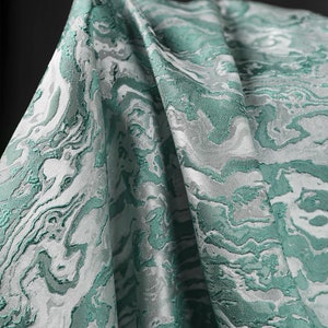 K&ouml;nnte beinhalten: Nahaufnahme eines gefalteten Stoffes mit einem marmorierten Muster in Teal und Wei&szlig;. Die Textur und die Details des Musters sind sichtbar.