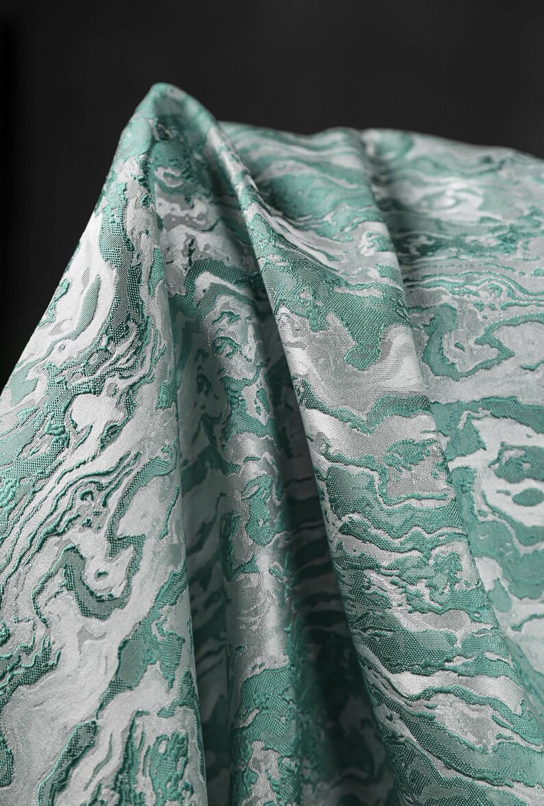 K&ouml;nnte beinhalten: Nahaufnahme eines gefalteten Stoffes mit einem marmorierten Muster in Teal und Wei&szlig;. Die Textur und die Details des Musters sind sichtbar.