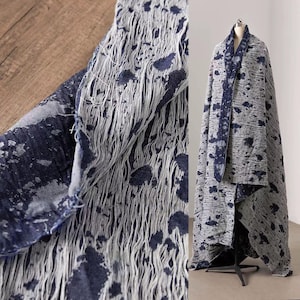 May include: A gray and navy blue textile with a unique, distressed design. The fabric features a pattern of dark blue splatters and vertical, frayed lines. The textile is draped over a mannequin, showcasing its full length and texture.