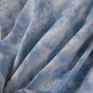Op de afbeelding: Close-up van een blauwe en witte tie-dye stof. De stof heeft een zachte, vloeiende textuur en een subtiele, vervaagde uitstraling.