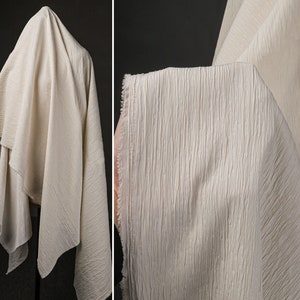 Può includere: Primo piano di un tessuto color crema con una texture grinzosa. Il tessuto è drappeggiato su una superficie e ha un aspetto morbido e fluido.