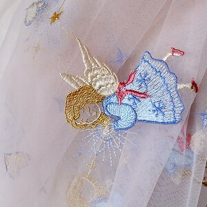 Puede incluir: Una delicada tela de tul blanco con detalles bordados de un &aacute;ngel alado en tonos azules, dorados y rosados. El &aacute;ngel est&aacute; representado con un vestido y alas flotantes, y la tela tiene un ligero brillo.