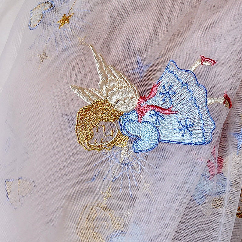 Puede incluir: Una delicada tela de tul blanco con detalles bordados de un &aacute;ngel alado en tonos azules, dorados y rosados. El &aacute;ngel est&aacute; representado con un vestido y alas flotantes, y la tela tiene un ligero brillo.