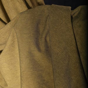 K&ouml;nnte beinhalten: Nahaufnahme eines olivgr&uuml;nen Stoffes mit dezentem Glanz. Der Stoff ist drapiert und zeigt Falten und Textur. Die Beleuchtung erzeugt Schatten und Glanzlichter, die die Oberfl&auml;che des Materials hervorheben.