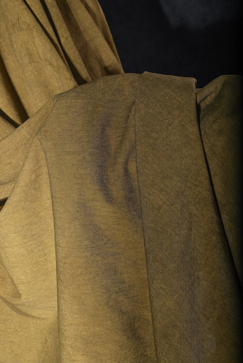 K&ouml;nnte beinhalten: Nahaufnahme eines olivgr&uuml;nen Stoffes mit dezentem Glanz. Der Stoff ist drapiert und zeigt Falten und Textur. Die Beleuchtung erzeugt Schatten und Glanzlichter, die die Oberfl&auml;che des Materials hervorheben.