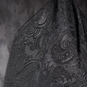K&ouml;nnte beinhalten: Nahaufnahme eines schwarzen Stoffes mit einem komplizierten, gepr&auml;gten Paisley-Muster. Der Stoff hat einen subtilen Glanz, und das Design weist wirbelnde, organische Formen auf. Das Material ist drapiert und erzeugt Schatten, die die Textur hervorheben.