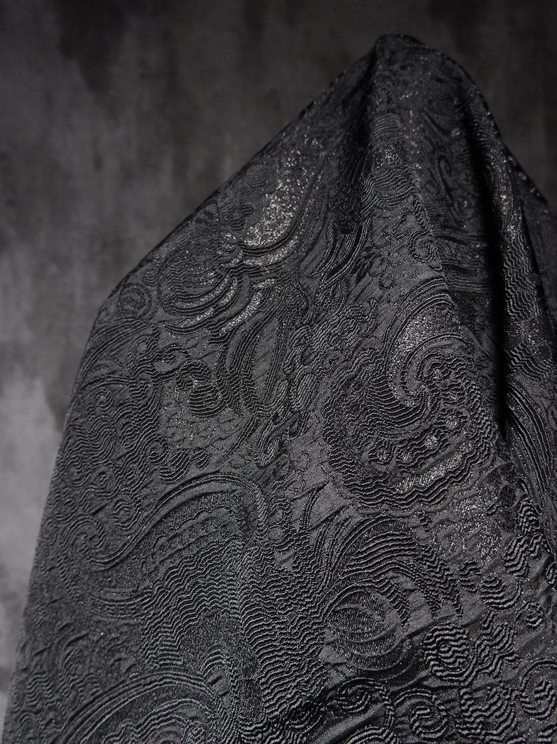 K&ouml;nnte beinhalten: Nahaufnahme eines schwarzen Stoffes mit einem komplizierten, gepr&auml;gten Paisley-Muster. Der Stoff hat einen subtilen Glanz, und das Design weist wirbelnde, organische Formen auf. Das Material ist drapiert und erzeugt Schatten, die die Textur hervorheben.