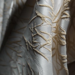 May include: Close-up of a silver fabric with a raised, textured pattern of beige lines and swirls.