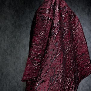 Op de afbeelding: Een gedrapeerd stuk stof in een diepe bordeauxrode kleur met een textuur die bijna vloeibaar lijkt. De stof heeft een subtiele glans en een patroon van donkere, onregelmatige lijnen, wat een luxueus en dramatisch effect creëert. De achtergrond is donkergrijs.