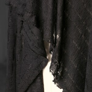 以下が含まれることがあります： 黒色のオープン織りの衣服のクローズアップ。ドレープのデザインで、生地は質感があり、縁はフリンジになっています。ショールまたはラップのようです。