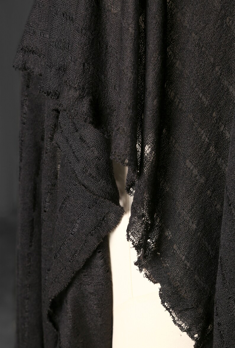 以下が含まれることがあります： 黒色のオープン織りの衣服のクローズアップ。ドレープのデザインで、生地は質感があり、縁はフリンジになっています。ショールまたはラップのようです。