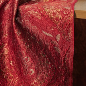 Może przedstawiać: Zbliżenie czerwonej tkaniny ze złotym wzorem paisley. Tkanina jest ułożona na drewnianej powierzchni, prezentując swoją fakturę i wzór. Bogaty czerwony kolor i metaliczne złote akcenty tworzą luksusową estetykę.