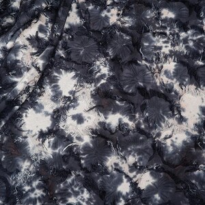 May include: Close-up of a fabric with a tie-dye pattern in shades of gray and white. The fabric has a textured appearance with frayed edges, creating a layered effect. The design features circular patterns resembling flowers.