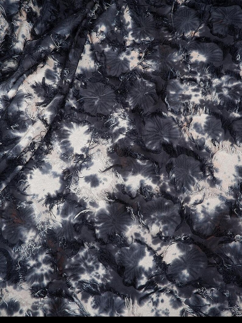 May include: Close-up of a fabric with a tie-dye pattern in shades of gray and white. The fabric has a textured appearance with frayed edges, creating a layered effect. The design features circular patterns resembling flowers.