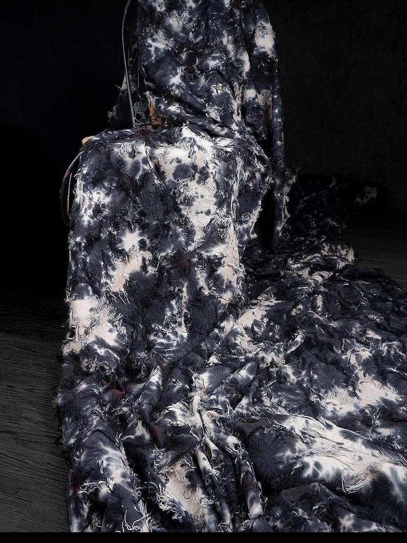 May include: A dark fabric with a tie-dye pattern in shades of black and white. The fabric is draped over a chair, with the edges frayed, creating a textured and layered effect. The overall aesthetic is modern and artistic.