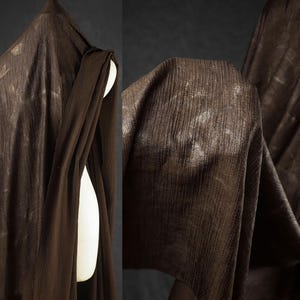 Peut inclure: Gros plan sur un tissu drapé brun foncé avec un motif texturé à côtes verticales. Le tissu est drapé sur un mannequin blanc, avec des plis et des ombres ajoutant de la profondeur. Le matériau semble léger et fluide.