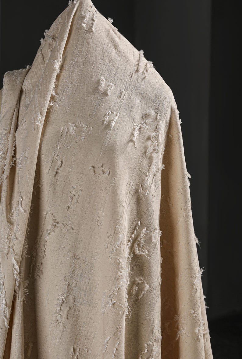 May include: A beige fabric with a textured, frayed surface. The fabric has a subtle pattern of small, raised bumps.