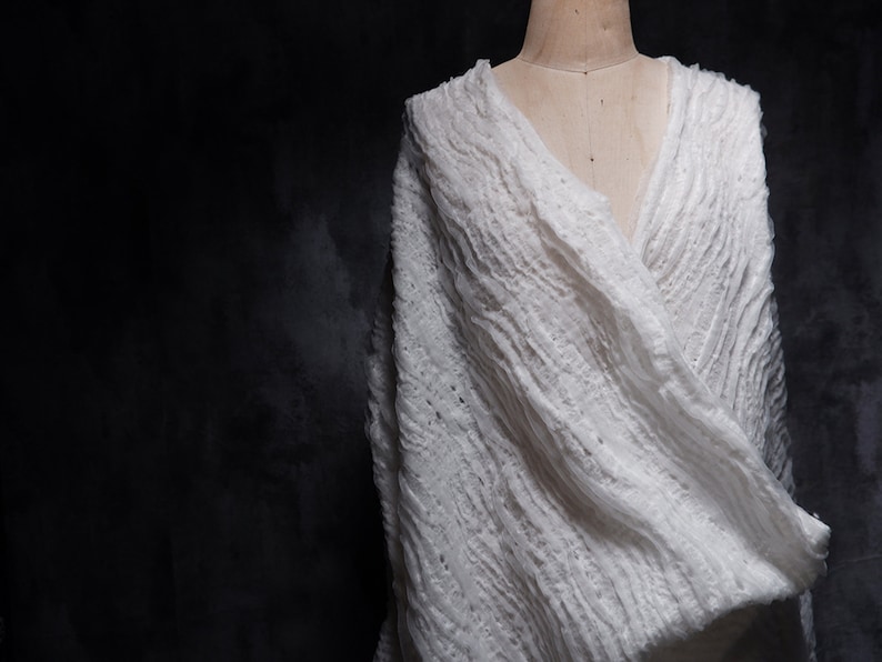 May include: A white, textured, crocheted shawl draped over a mannequin. The shawl has a loose, flowing design and is made of a soft, lightweight fabric.