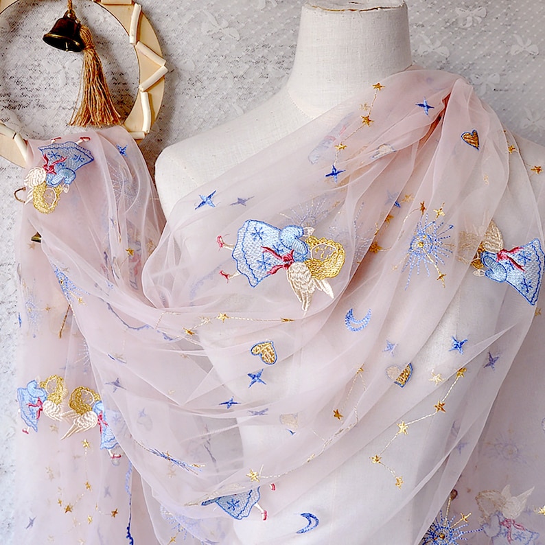 Puede incluir: Tela de tul rosa con &aacute;ngeles, estrellas y lunas bordadas en oro y azul. La tela es transparente y delicada.