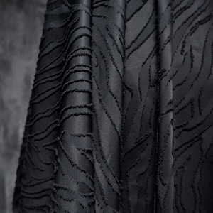Pode incluir: Close-up de um tecido preto drapeado com um padrão texturizado em relevo. O tecido tem um brilho acetinado e um design de elementos lineares curvos. A textura é criada com um ponto em relevo e com franjas, adicionando profundidade.
