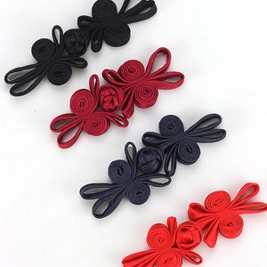 Può includere: Cinque paia di bottoni decorativi in tessuto di colore nero, rosso e blu scuro. I bottoni hanno la forma di un fiore con un centro annodato.