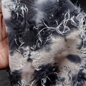 May include: Close-up of a distressed fabric with a tie-dye pattern in shades of gray and black. The edges of the fabric are frayed, with white and black threads creating a textured effect. The fabric appears to be soft and lightweight.