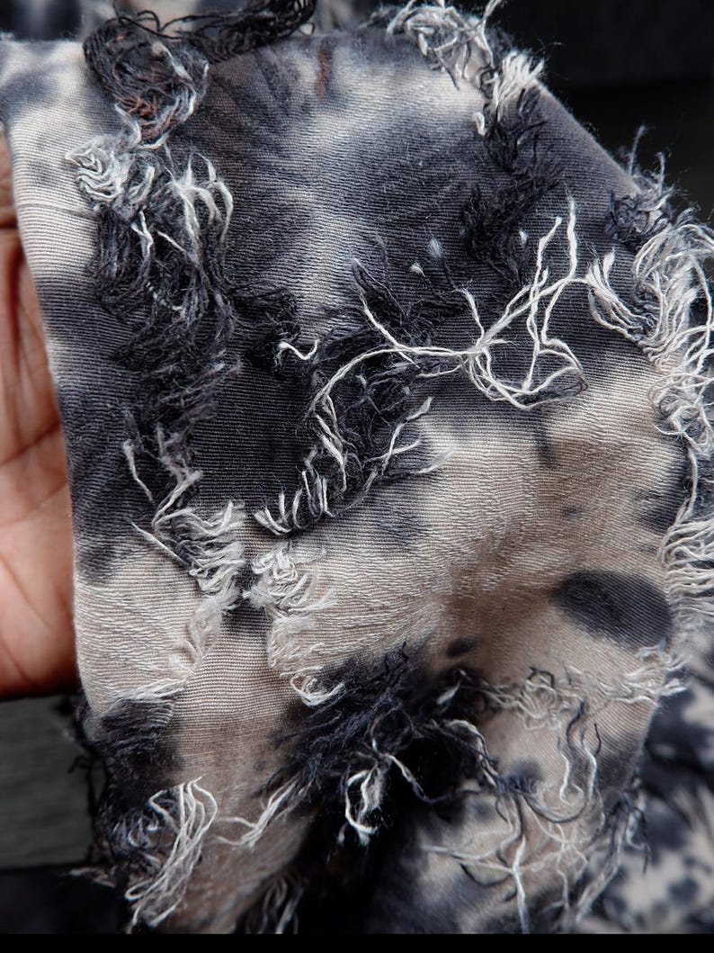 May include: Close-up of a distressed fabric with a tie-dye pattern in shades of gray and black. The edges of the fabric are frayed, with white and black threads creating a textured effect. The fabric appears to be soft and lightweight.