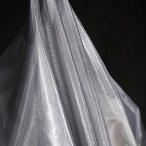 Może przedstawiać: Drapowana, połyskująca srebrna tkanina na ciemnym tle. Materiał ma delikatną, przezroczystą jakość, z widoczną tkaną fakturą i fałdami, kt&oacute;re chwytają światło, tworząc refleksy i cienie. Tkanina wydaje się lekka i zwiewna.