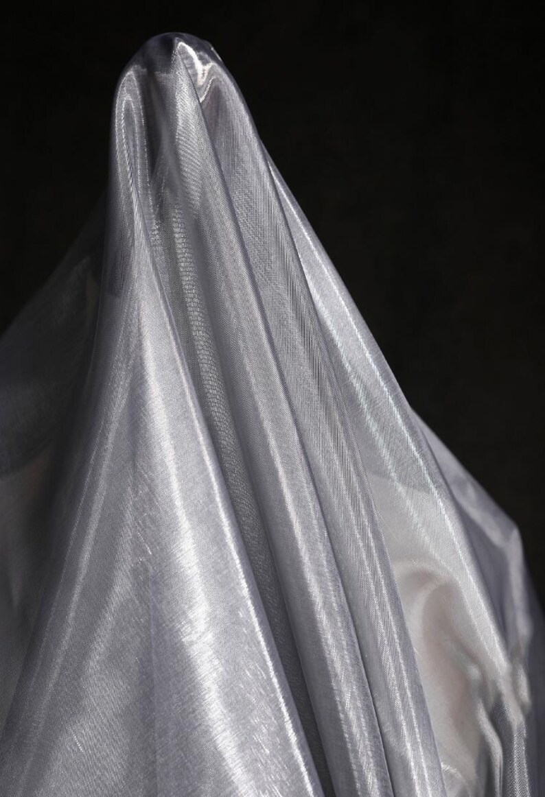 Może przedstawiać: Drapowana, połyskująca srebrna tkanina na ciemnym tle. Materiał ma delikatną, przezroczystą jakość, z widoczną tkaną fakturą i fałdami, kt&oacute;re chwytają światło, tworząc refleksy i cienie. Tkanina wydaje się lekka i zwiewna.