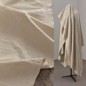 May include: Off-white, textured fabric with a reptile-like pattern. The fabric is draped over a surface and also draped over a mannequin. The fabric has a subtle sheen and a detailed, scale-like texture, suggesting a luxurious material.