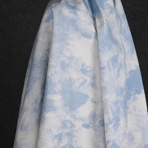 Op de afbeelding: Een witte stof met een blauw wolkenachtig patroon. De stof is gevouwen en over een oppervlak gelegd.