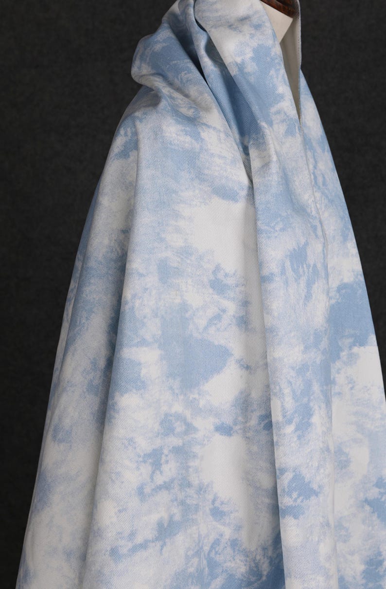 Op de afbeelding: Een witte stof met een blauw wolkenachtig patroon. De stof is gevouwen en over een oppervlak gelegd.
