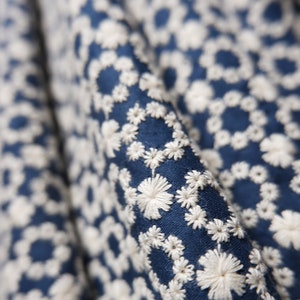 May include: Close-up of a blue fabric with a white embroidered floral pattern. The fabric is textured and has a subtle sheen.