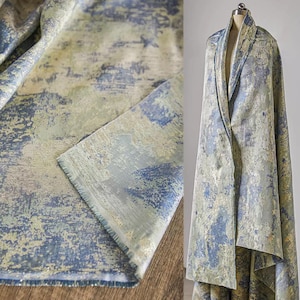 May include: Textile fabric with a distressed, abstract design in shades of blue, gray, and gold. The fabric is draped over a mannequin and also shown folded, revealing the texture and color variations. The fabric has a soft, flowing appearance.